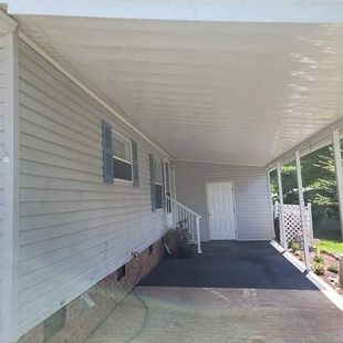 A light gray mobile home with a covered porch, white columns, and a dark asphalt driveway.