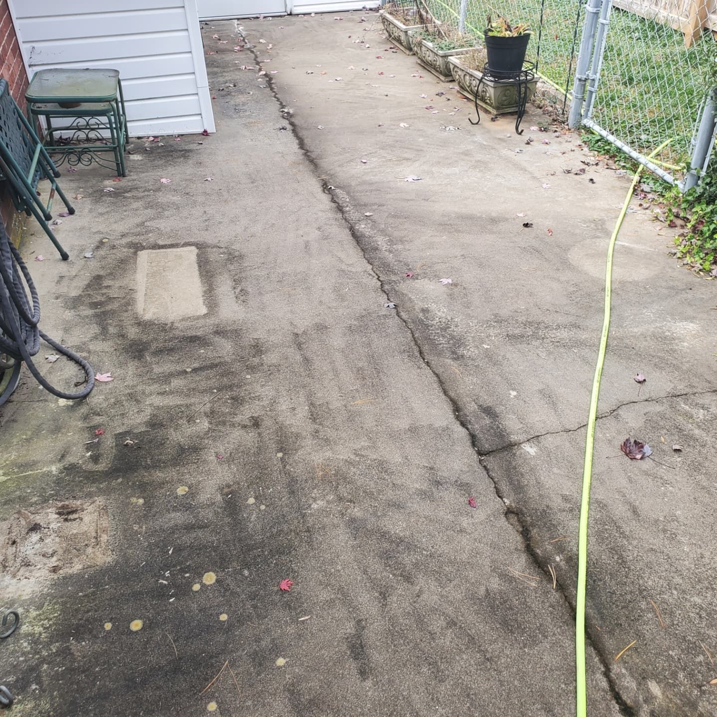 Concrete walkway in an outdoor space, with cracks and discoloration, next to a white building, fence, and hose.