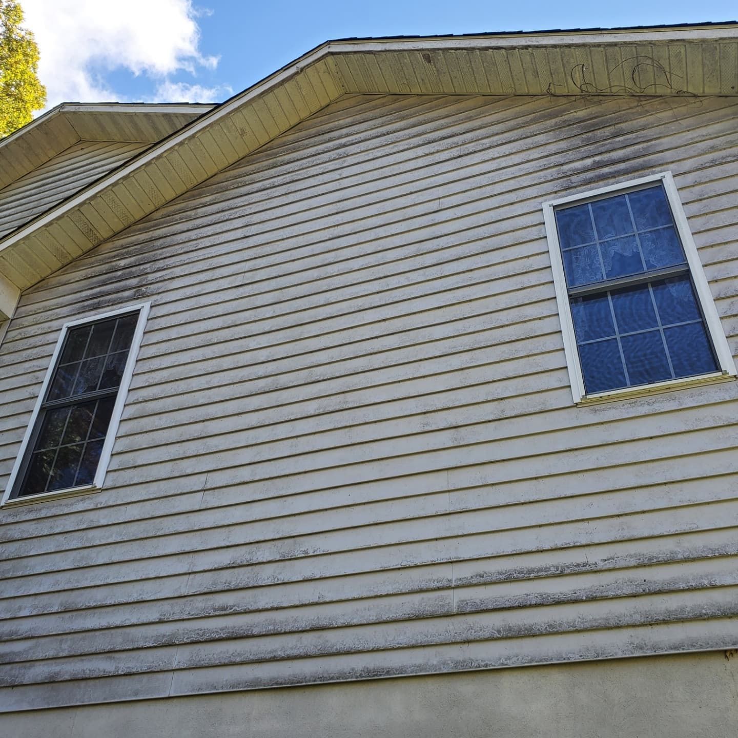 Side of a house with two windows and weathered, gray siding under a partly cloudy sky.
