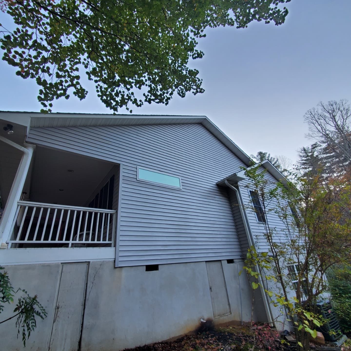 Gray-sided house with a porch and a light blue rectangle on the wall, trees overhead.