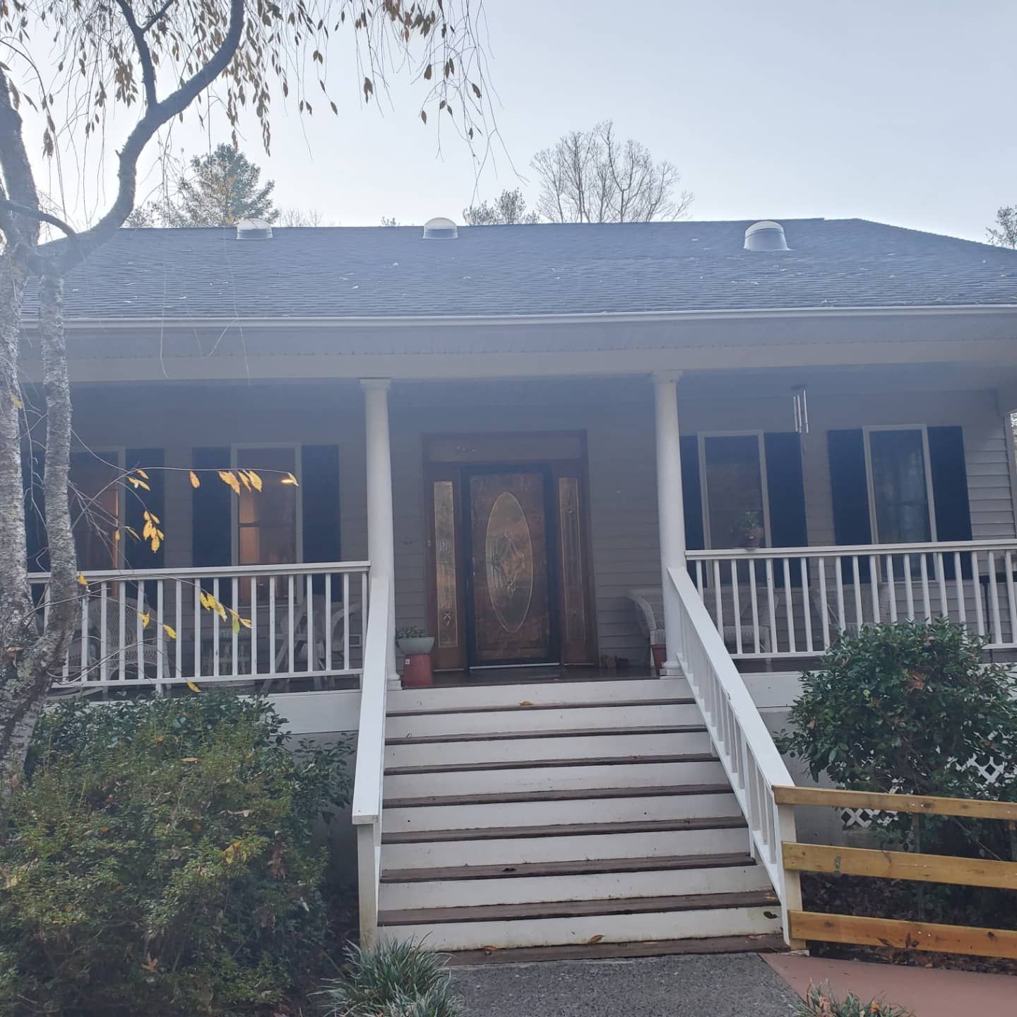 House with white porch, steps, and dark door. Gray roof. Trees and bushes in front.