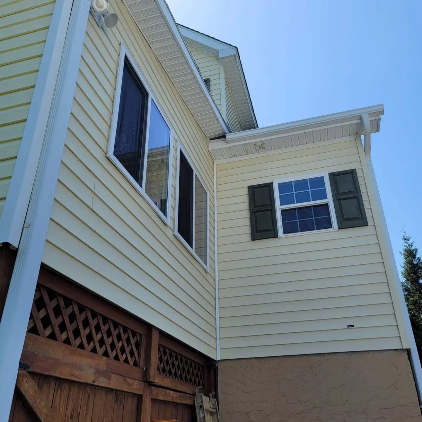 Yellow house exterior with windows, green shutters, and brown lattice detail, against a blue sky.