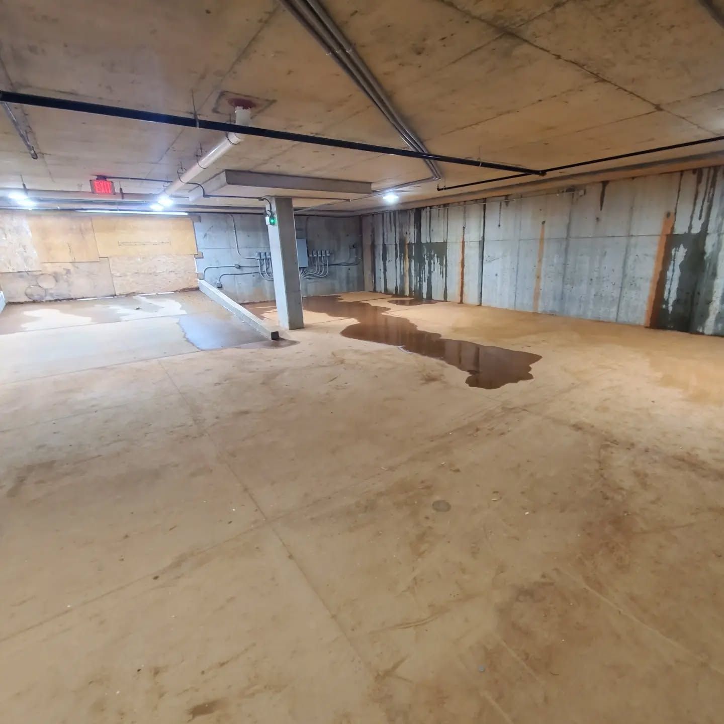 Water pools on the floor of a concrete basement. Walls and ceiling show water stains.