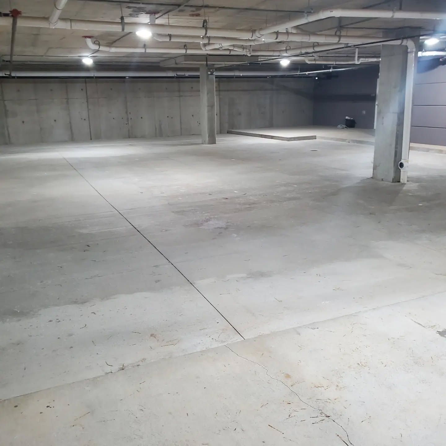 Empty concrete basement with support columns, pipes overhead, and ceiling lights.