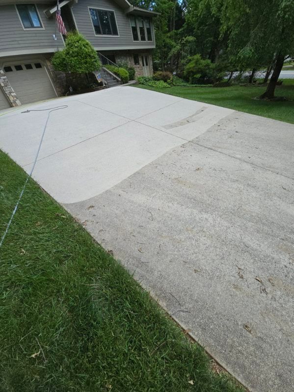 Driveway, half-cleaned, transitioning from dirty to light gray concrete, next to green grass and a house.