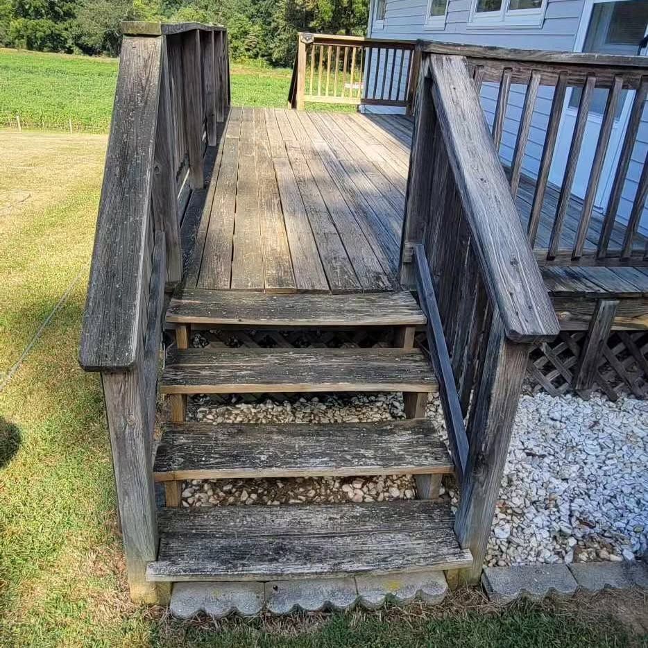 Wooden deck with steps, weathered gray wood. Gravel base, railing. Sunny outdoors.
