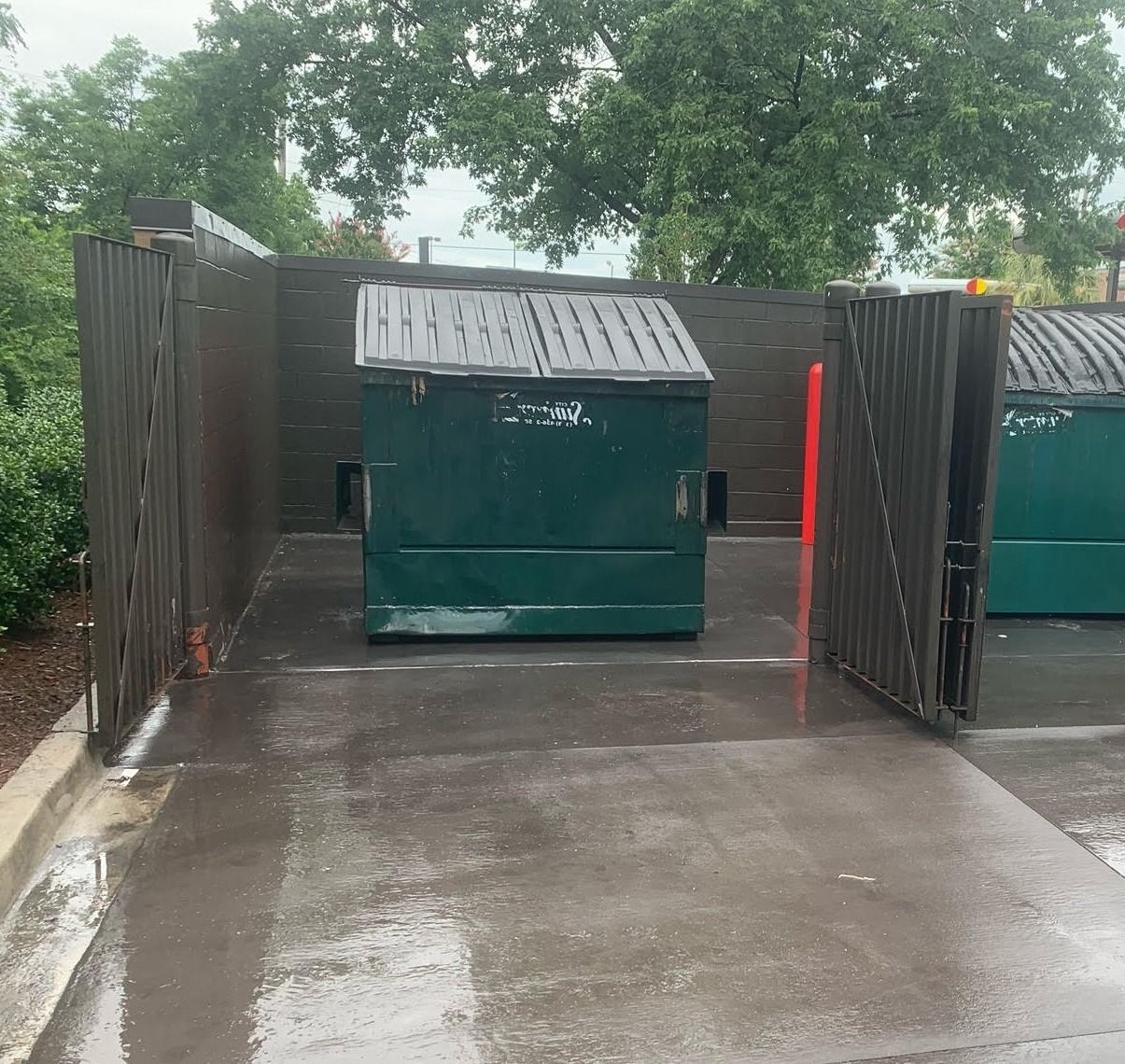 Green dumpsters in a concrete enclosure with wooden fencing, wet ground.