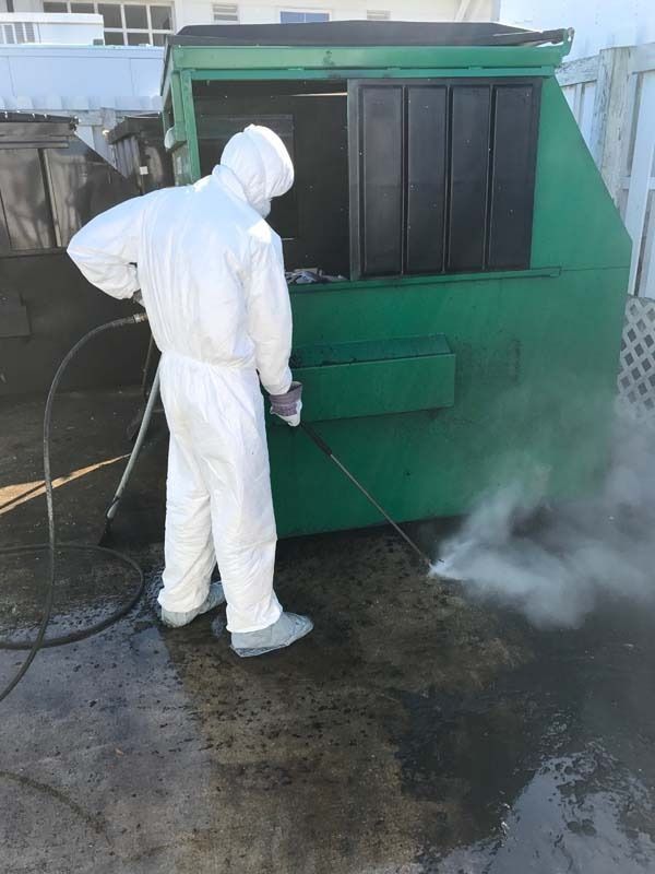 Person in white protective suit power washing a green dumpster outside.
