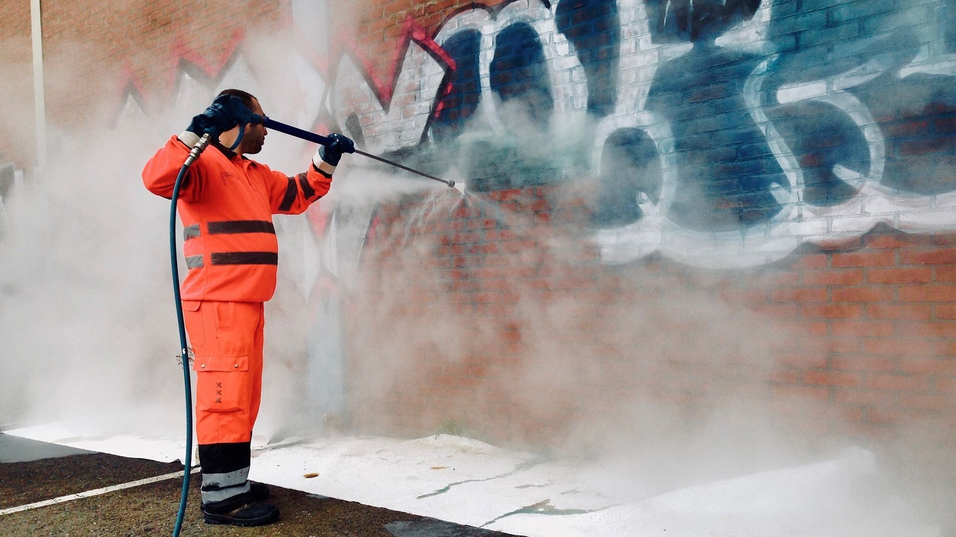 Person in orange suit power washing graffiti from brick wall, spraying water.