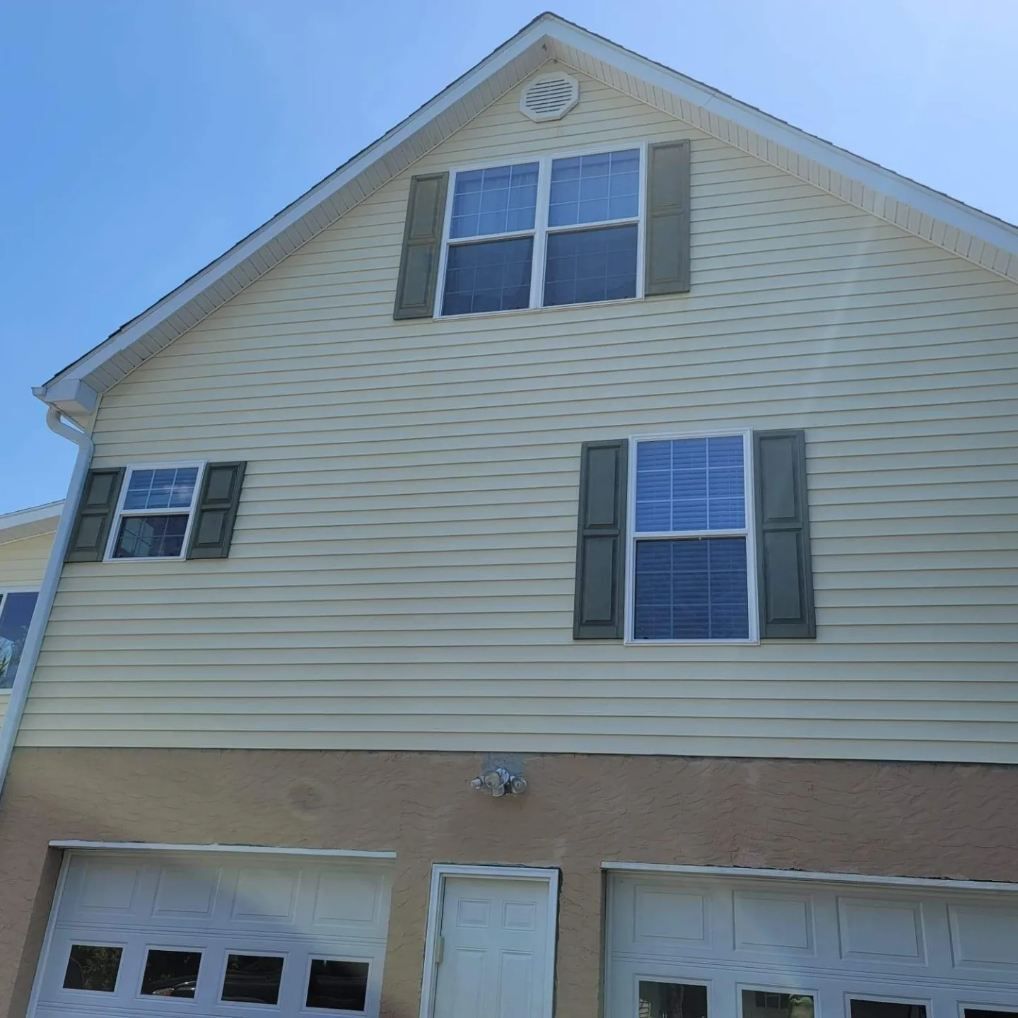Two-story house with tan siding, green shutters, white garage doors, and a blue sky.