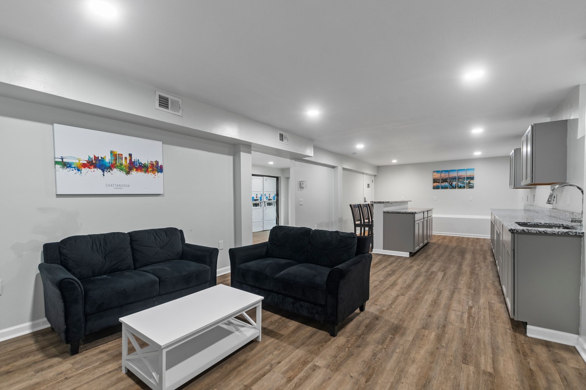 A modern living room and kitchen open-concept area featuring two black sofas, a white coffee table, and gray cabinetry.