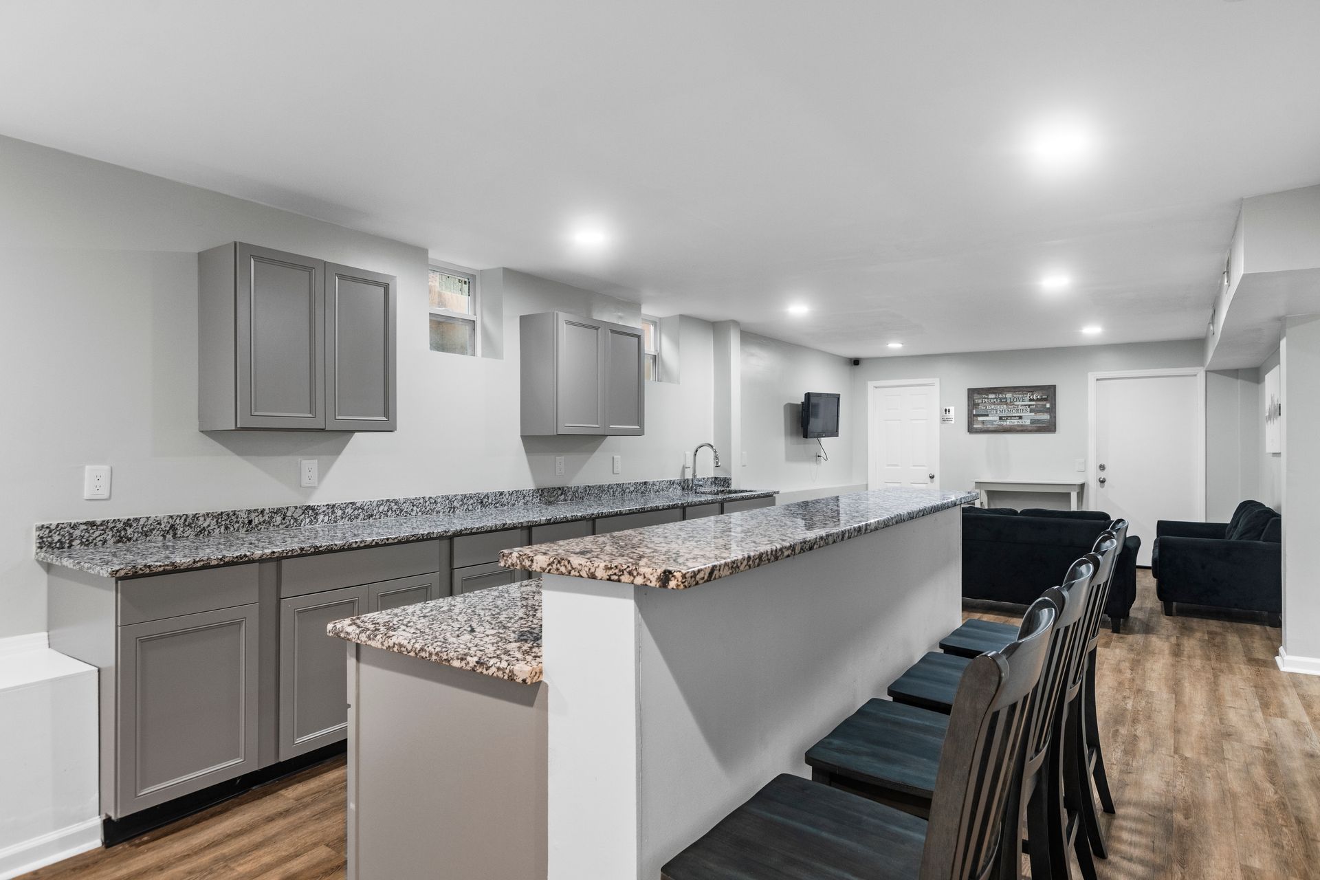 A basement wet bar featuring gray cabinets, speckled granite countertops, and a peninsula with bar stools.