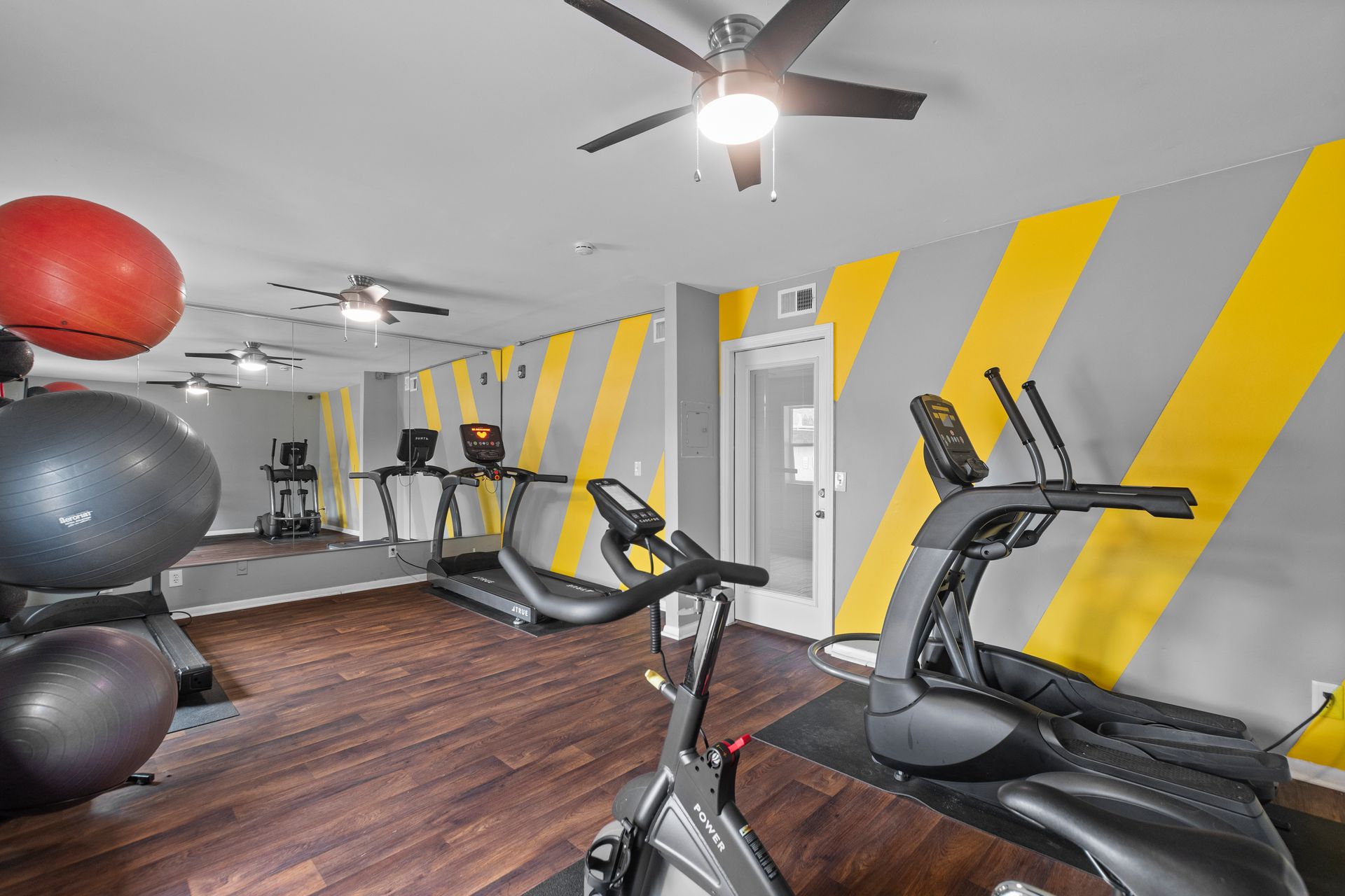 A small gym with exercise bikes and a treadmill, featuring grey and yellow diagonal striped walls and wooden floors.