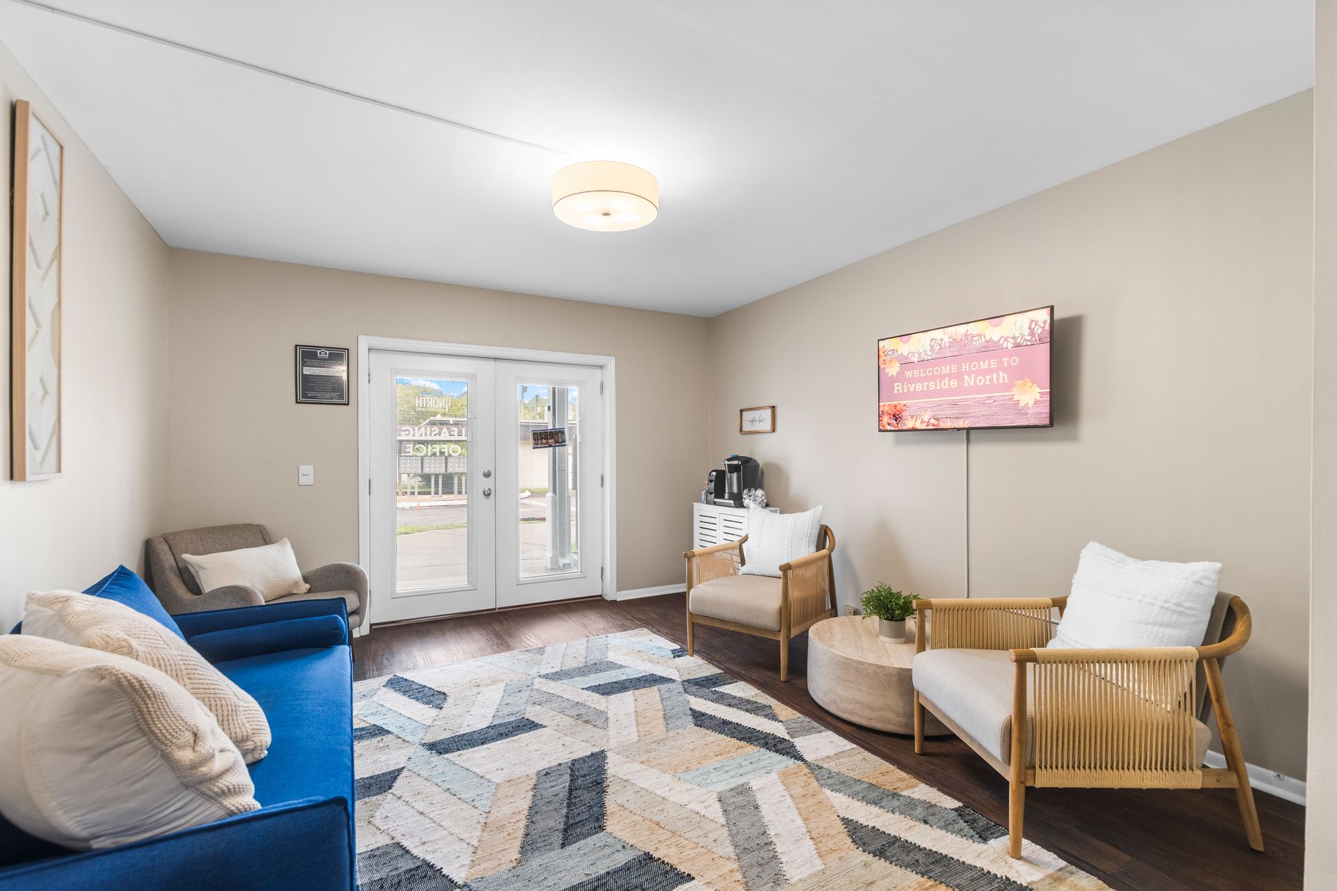 A bright waiting room with a blue sofa, two armchairs, a patterned rug, and a wall-mounted TV by glass double doors.