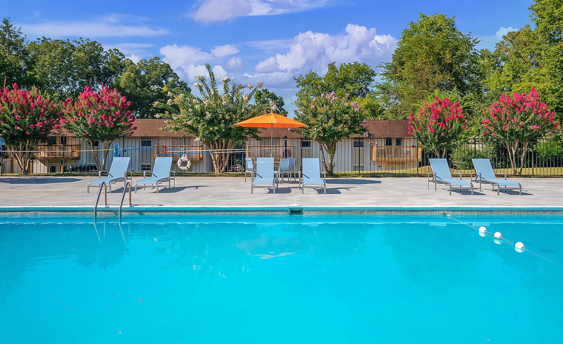 A bright blue swimming pool sits in front of a row of small cabins with blooming pink trees and an orange umbrella.