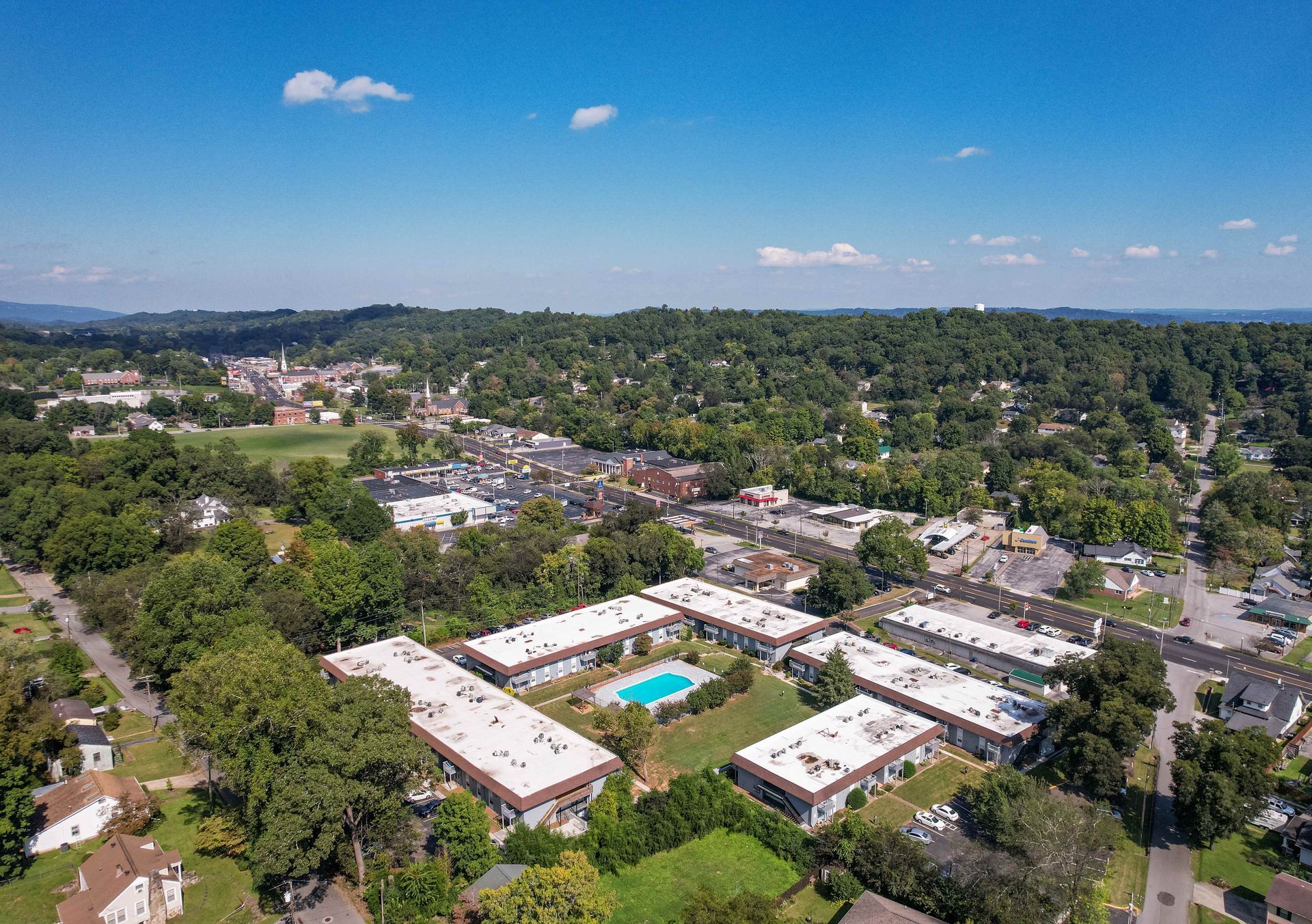 Aerial view of an apartment complex with a central swimming pool, surrounded by trees and a town under a clear blue sky.