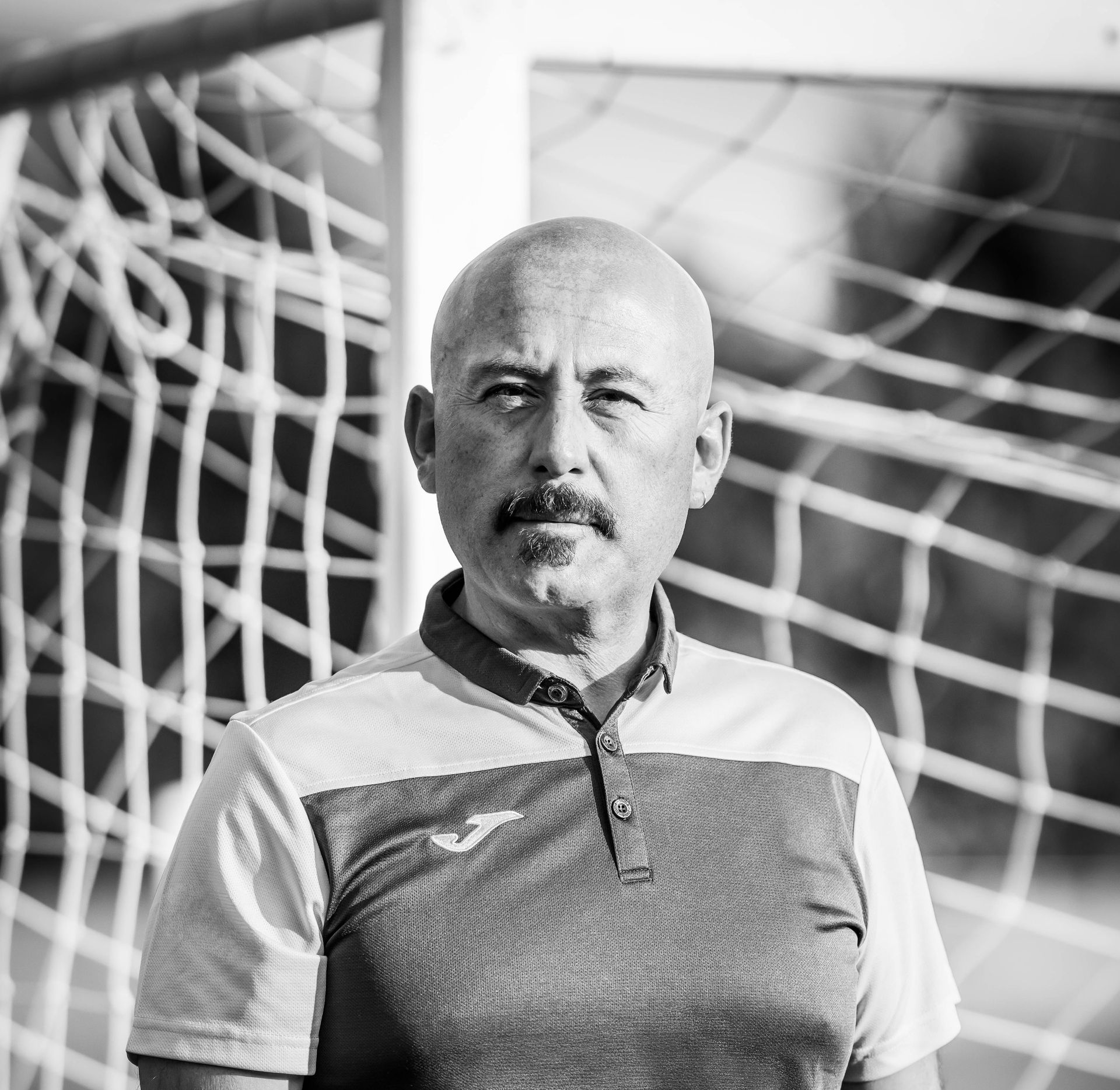 Bald man with mustache, wearing a polo shirt, stands in front of a soccer net.