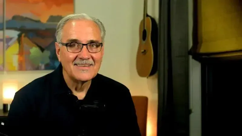 A man wearing glasses and a mustache is sitting in front of a guitar.
