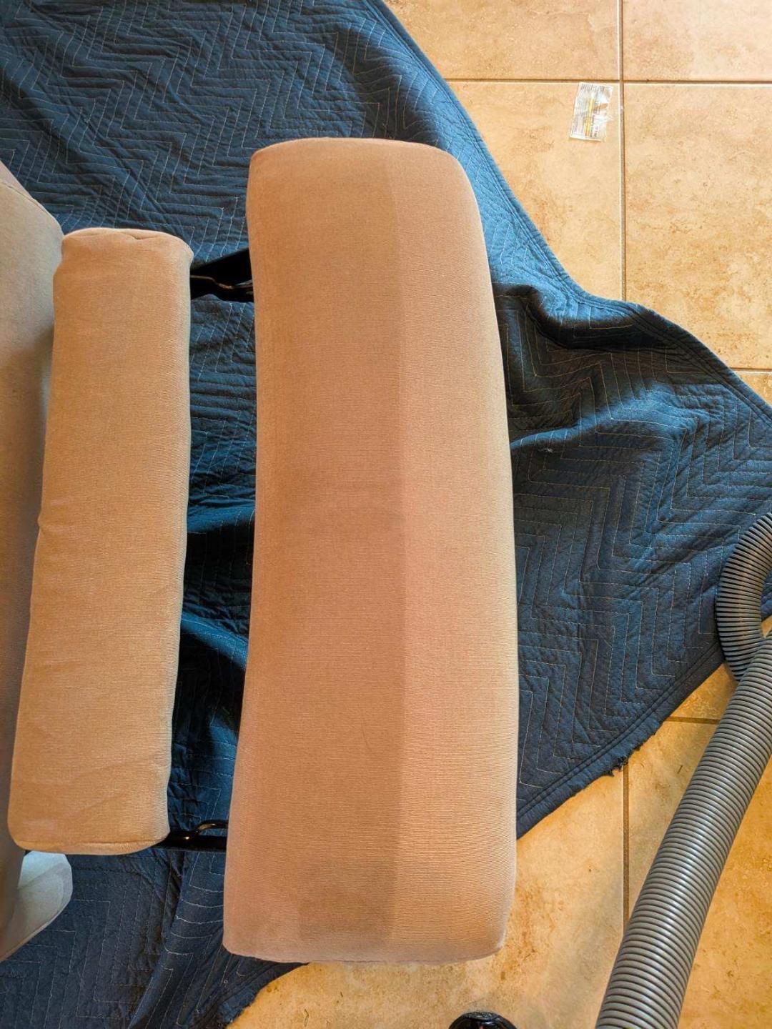 Two tan chair arms on a blue moving blanket, next to a vacuum hose, on a tile floor.