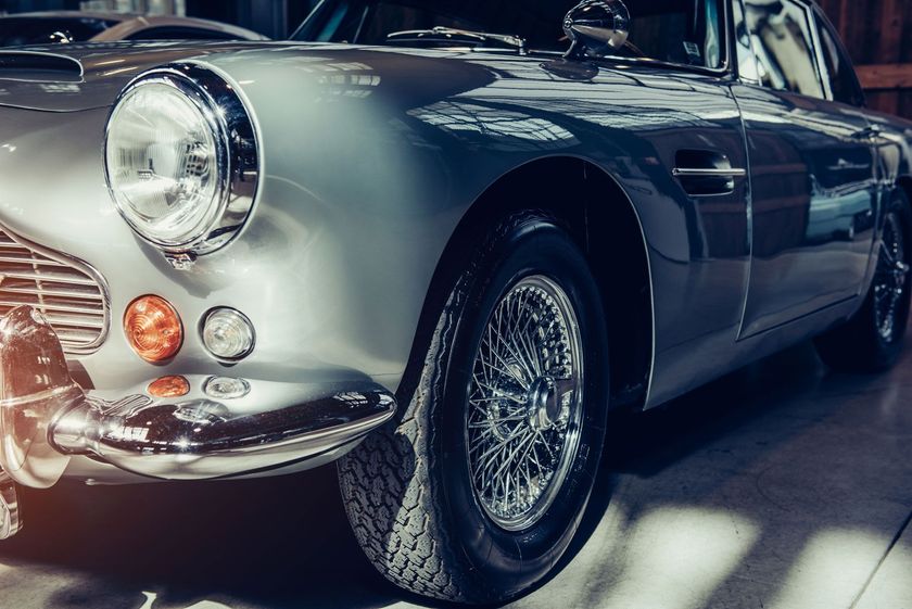 Silver vintage sports car with wire wheels, headlight on, parked indoors.