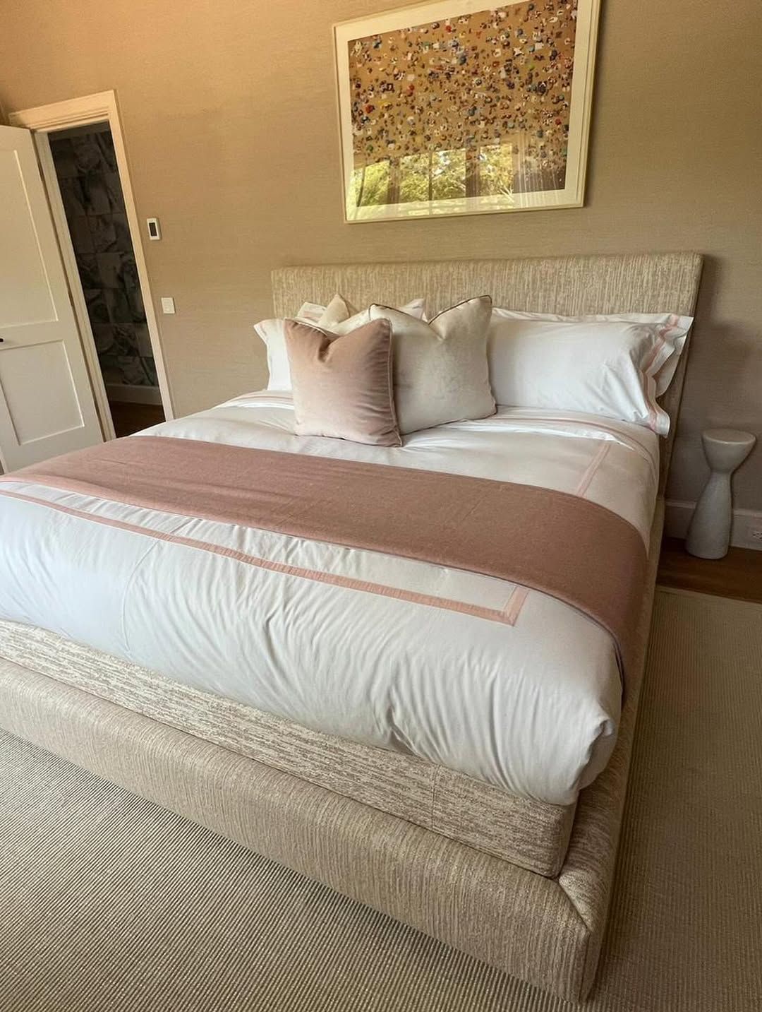 Beige bed with white bedding and pink accents. Abstract art on wall, next to a door.