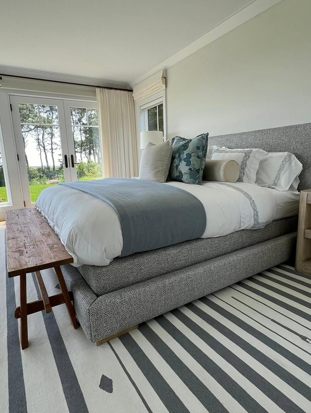 Bedroom with bed, bench, and striped rug, near glass doors overlooking greenery.