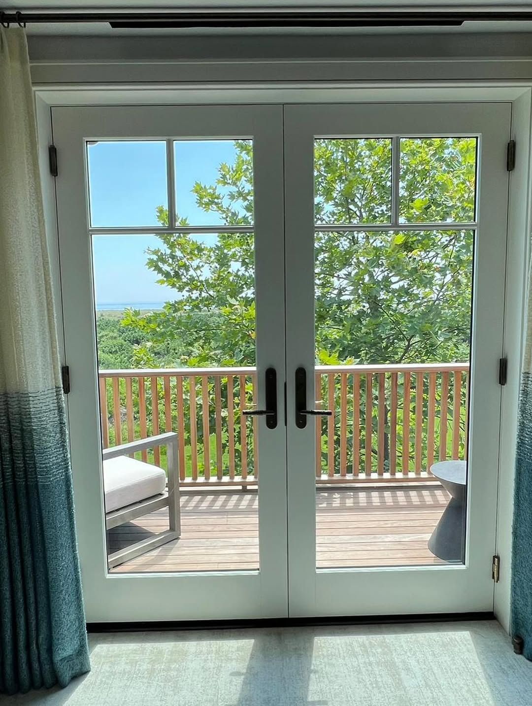 French doors open to a deck with greenery and a partial water view. Curtains frame the doors.