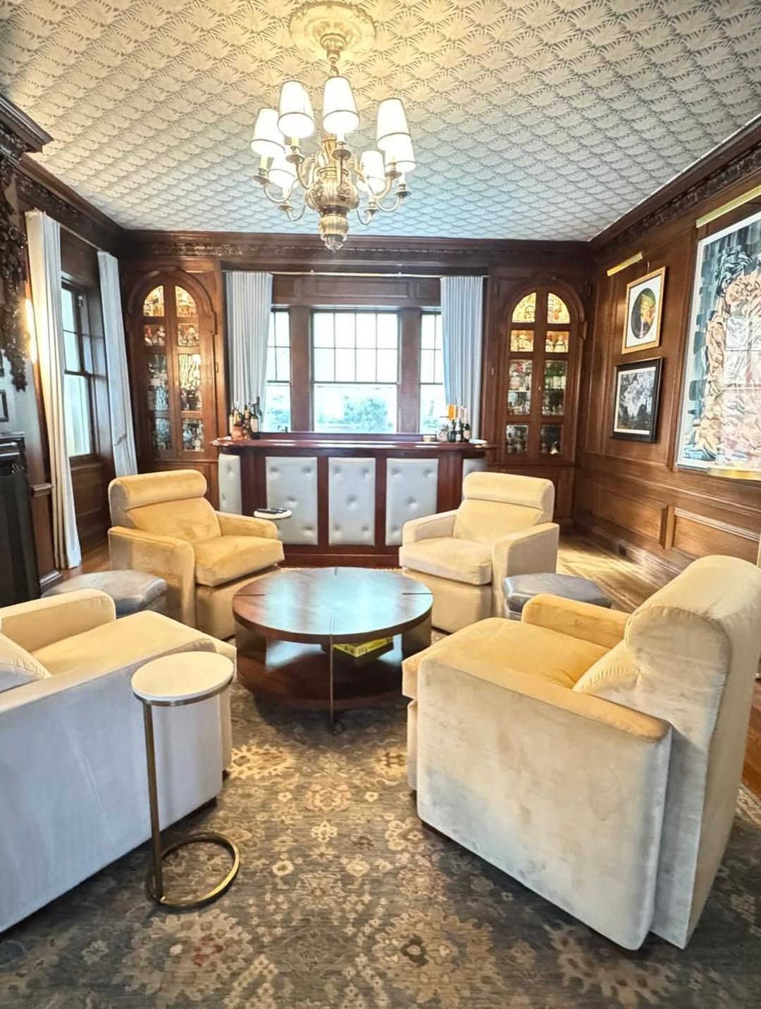 Cozy room with cream armchairs around a coffee table, built-in bar, wood paneling, ornate ceiling, chandelier.