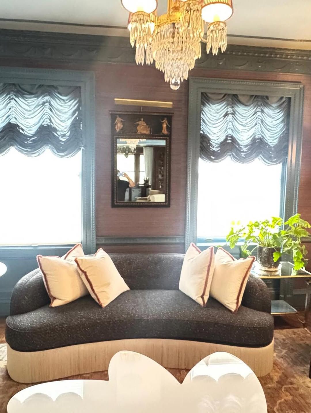 Living room with dark patterned sofa, white pillows, ornate chandelier, windows with blue drapes, and a decorative mirror.