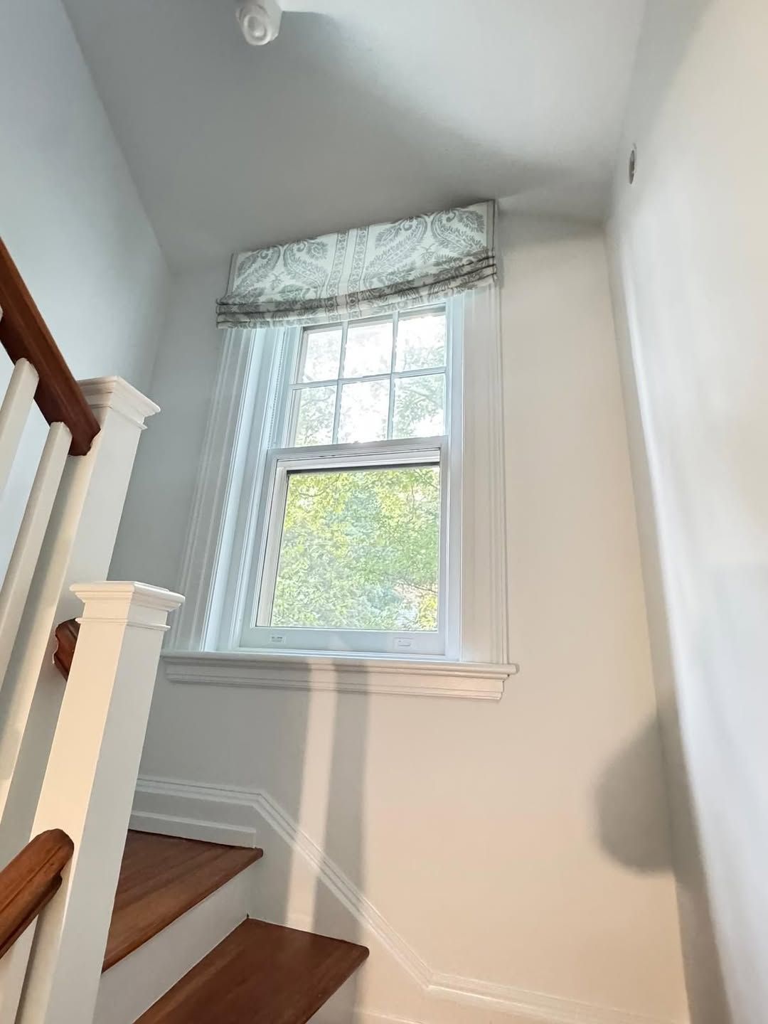 Stairwell window with patterned valance, sheer curtain, and wooden stairs with white trim.