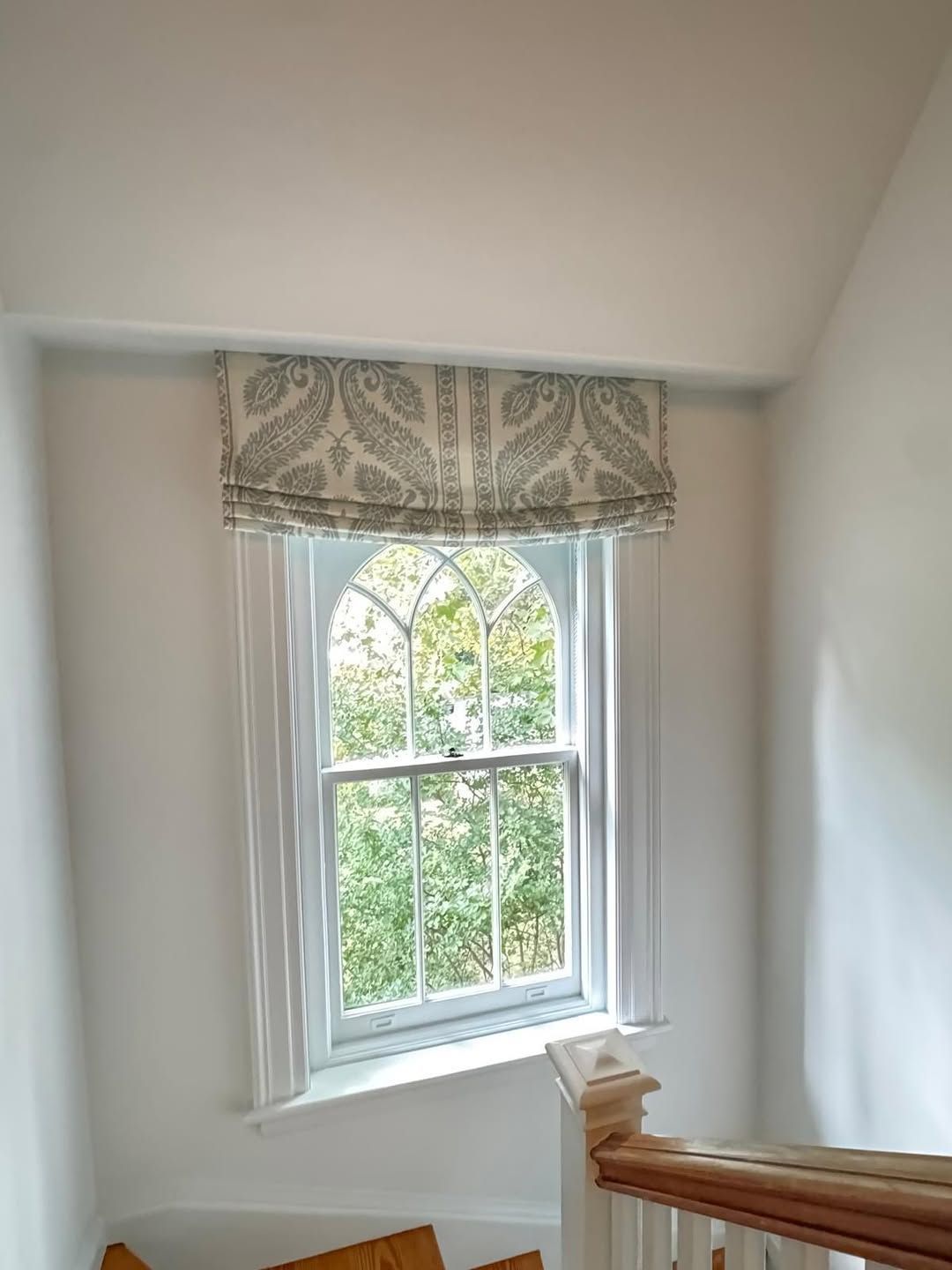 Window with arched top, white frame, paisley roman shade. Sunlight streams through glass.