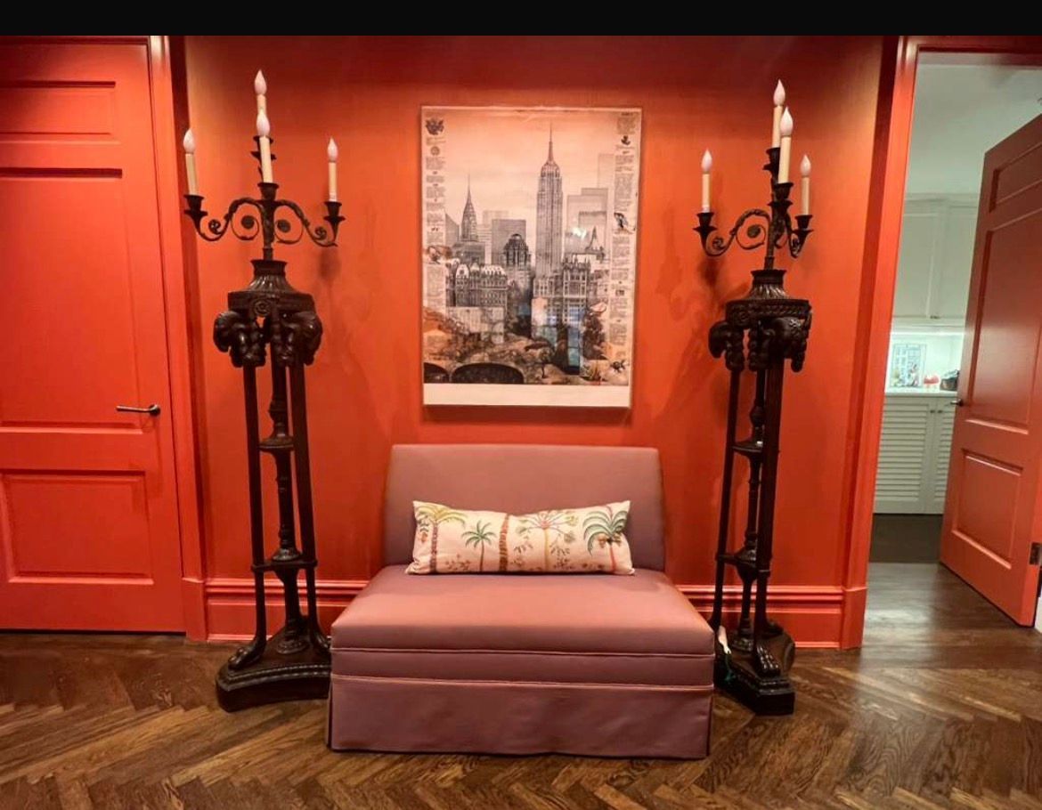 An entryway with orange walls, ornate black candle holders, art, and a small mauve bench.