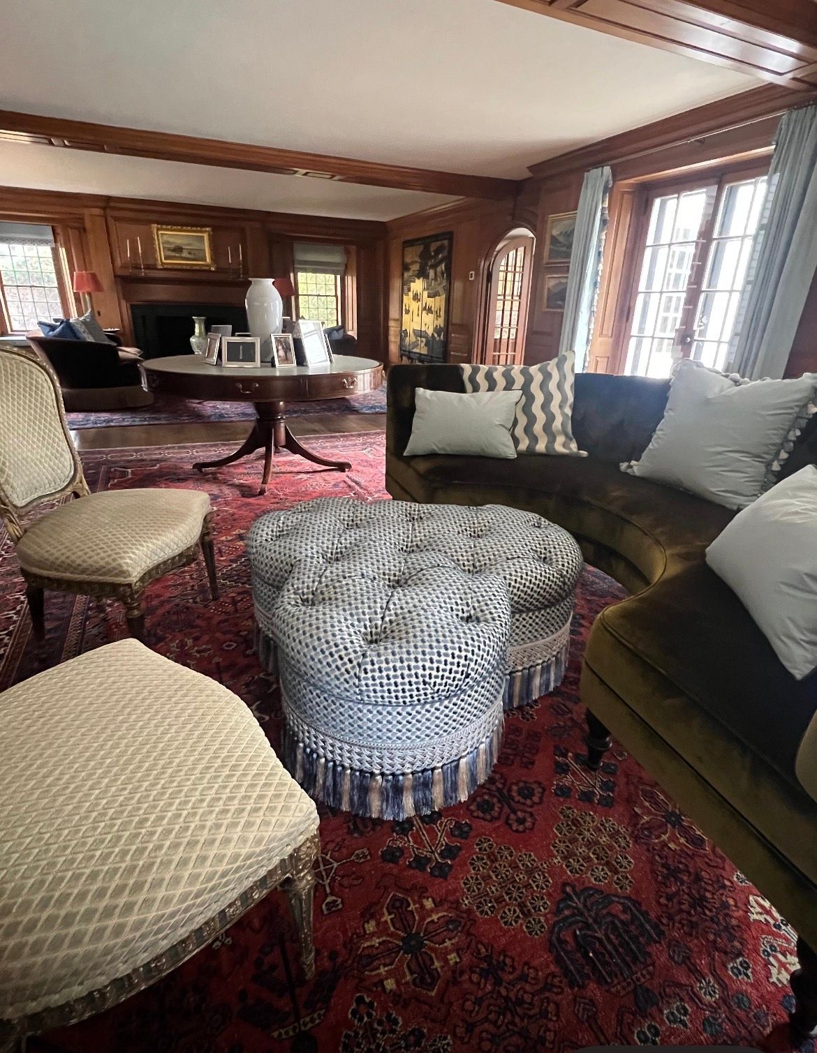 Living room with tufted ottoman, velvet sofa, antique chairs, and oriental rug. Wooden beams and paneling.