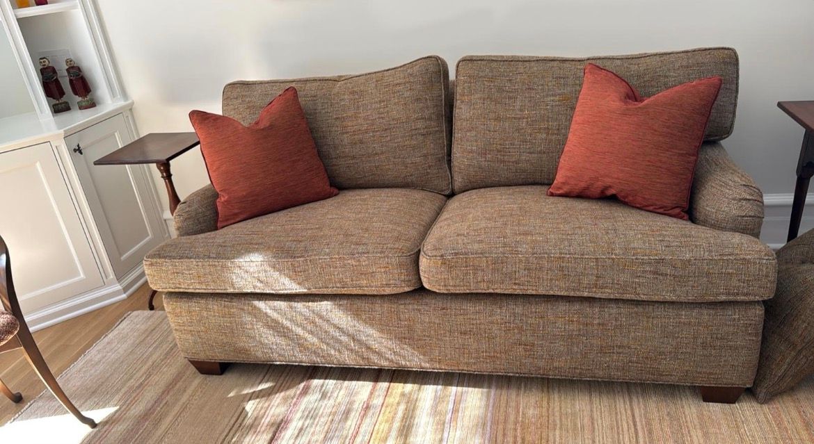 Brown tweed loveseat with two orange pillows, two small side tables, and a rug in a bright room.
