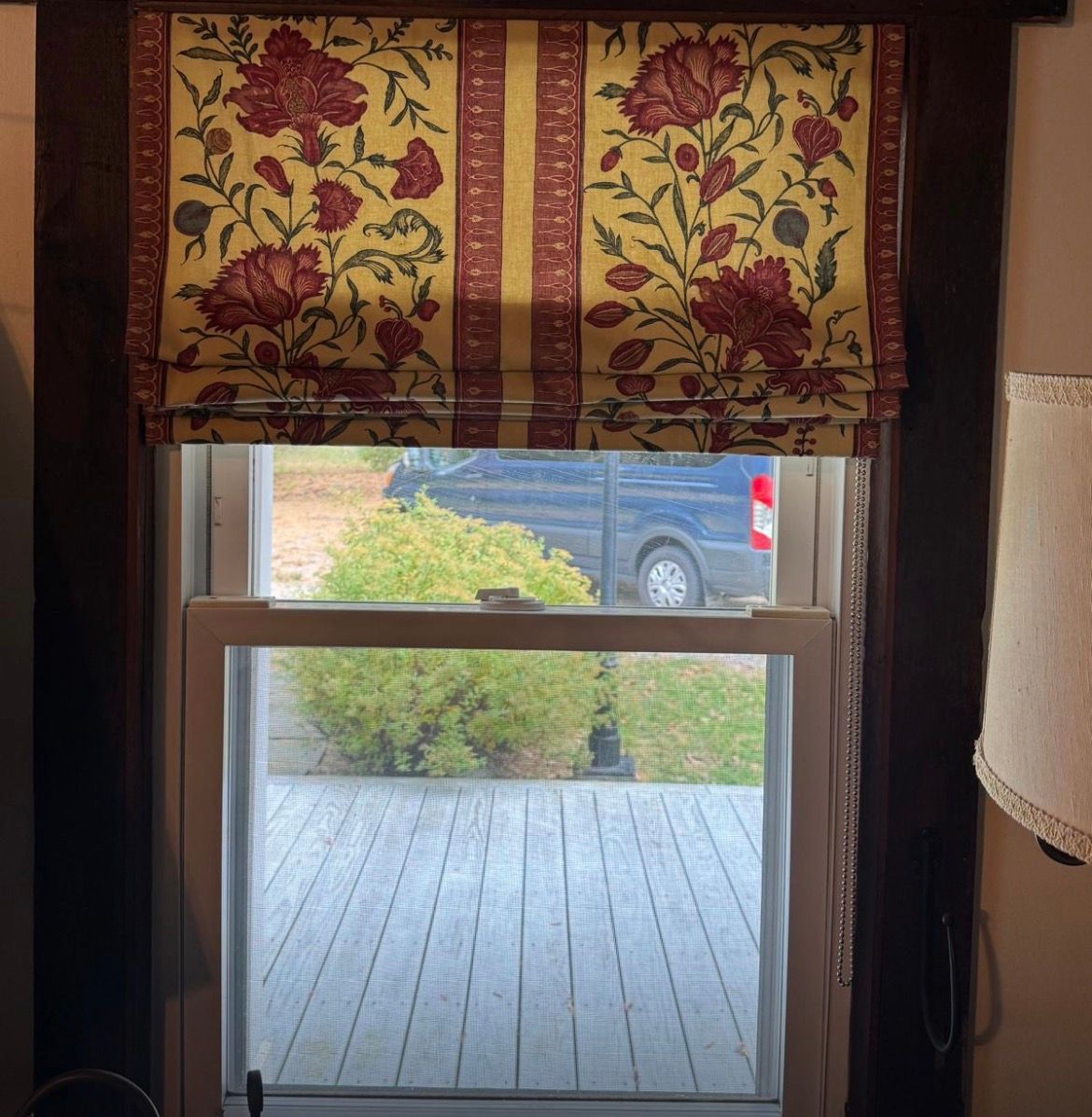 Window with a floral patterned Roman shade, revealing an outdoor scene with a deck, bushes, and a van.