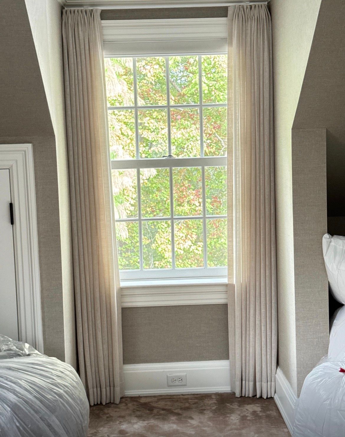 Tall window with beige drapes, green tree visible through the glass. Room with textured walls and rug.