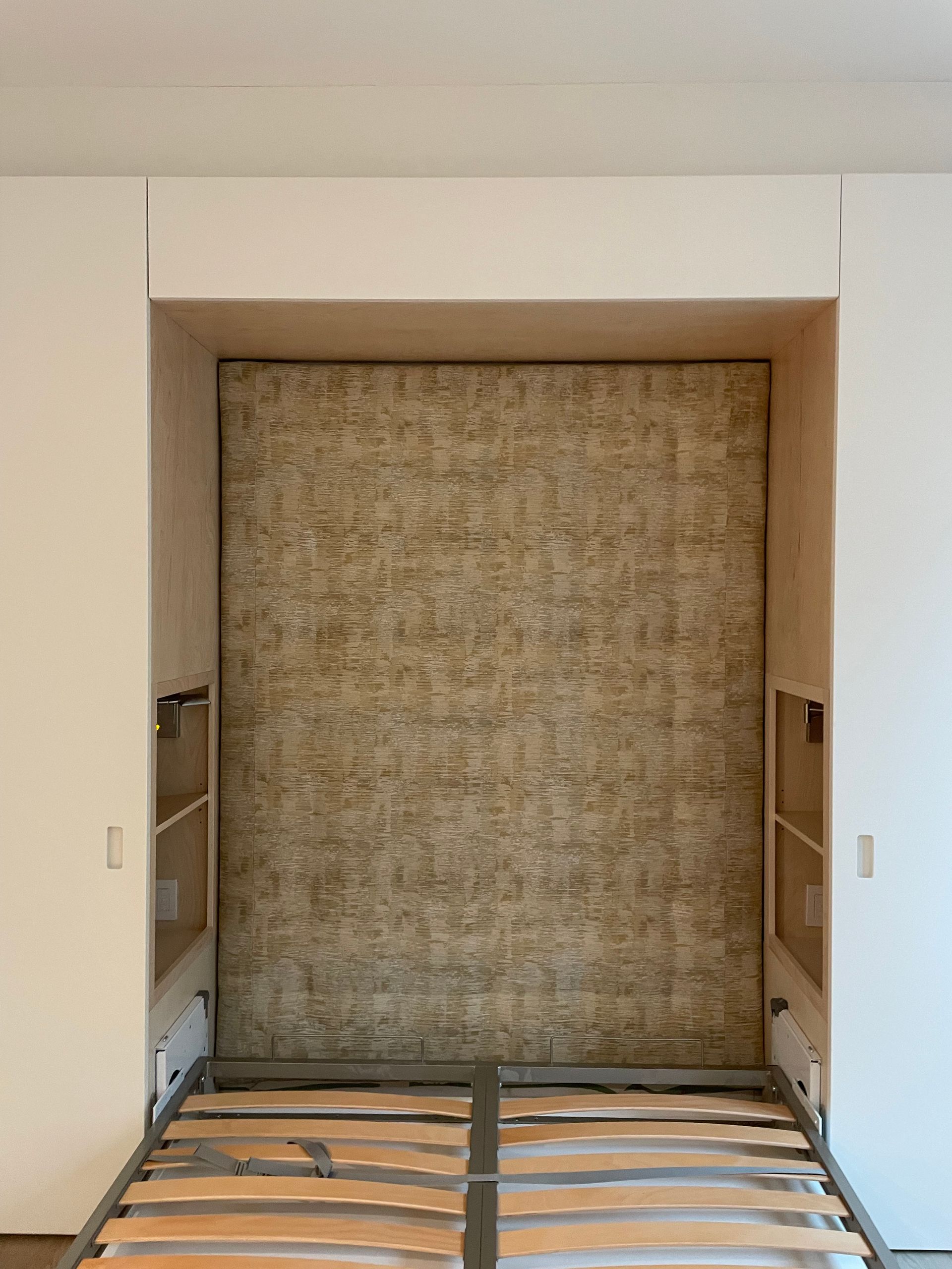 A wall bed in a recess, with shelves on either side. The bed's frame is visible, and the headboard is fabric-covered.