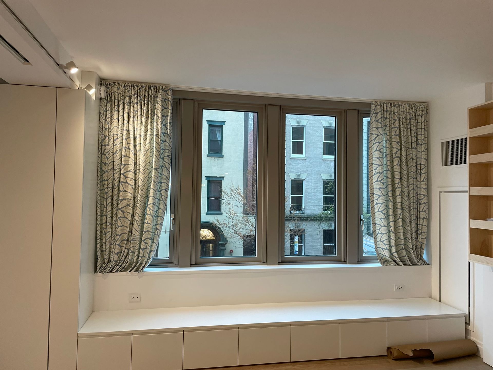 Window seat with a view of a city building, gray-framed windows, and patterned curtains.