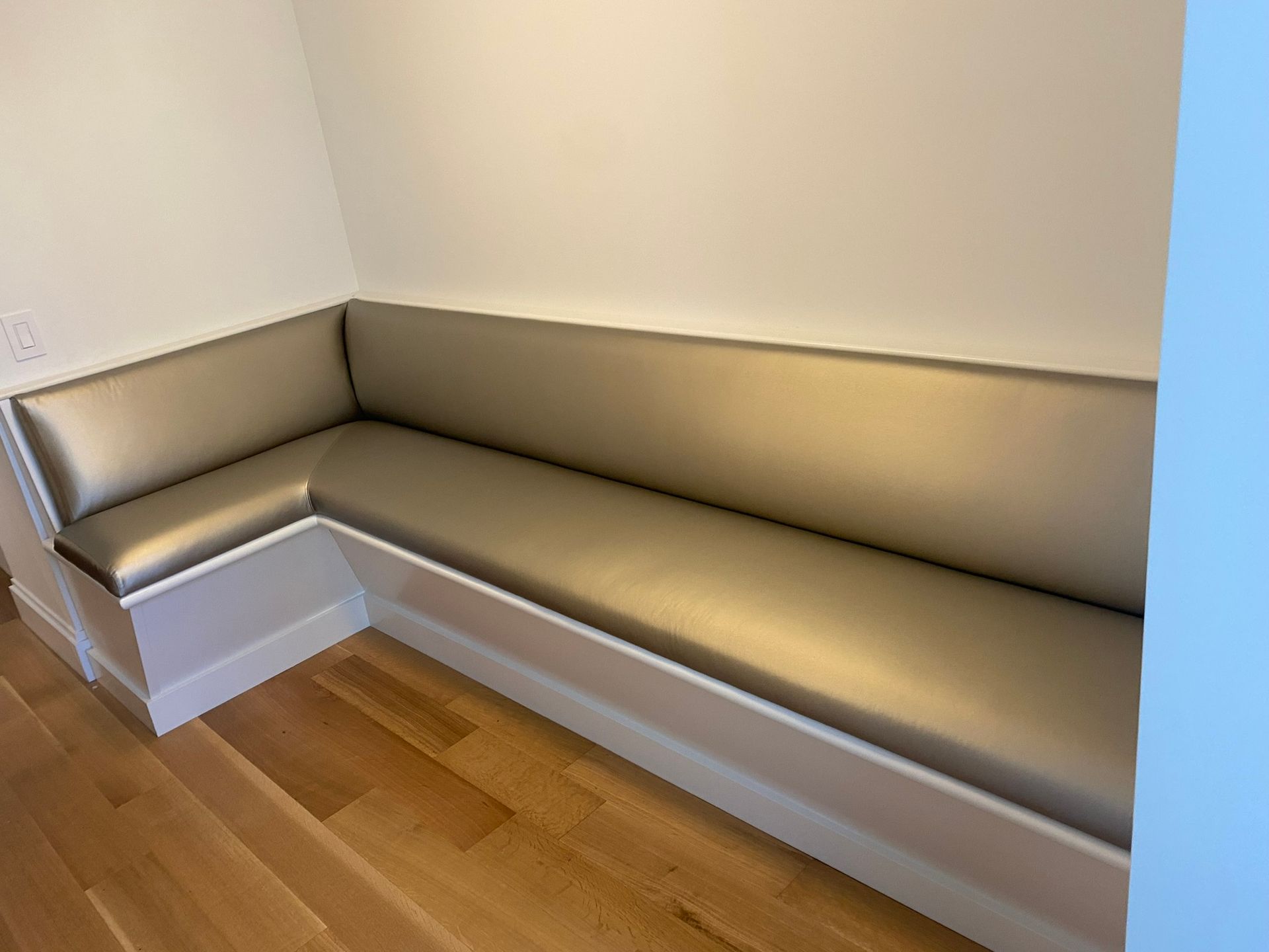Built-in, corner bench with light wood floors and white trim, against off-white walls.