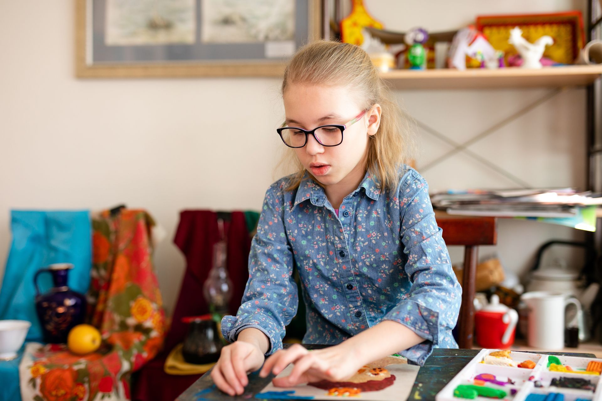 A young girl wearing glasses is sitting at a table painting a picture.