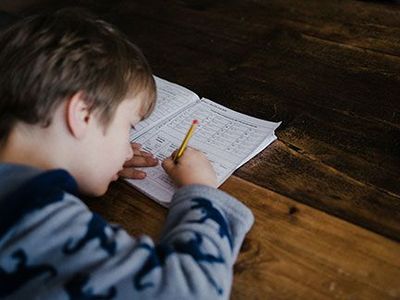 A young boy is sitting at a table writing in a notebook with a pencil.