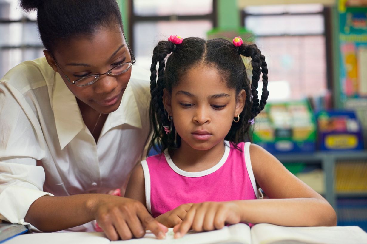 A woman is helping a little girl read a book in a classroom.