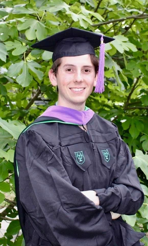 A young man wearing a graduation cap and gown is standing with his arms crossed.