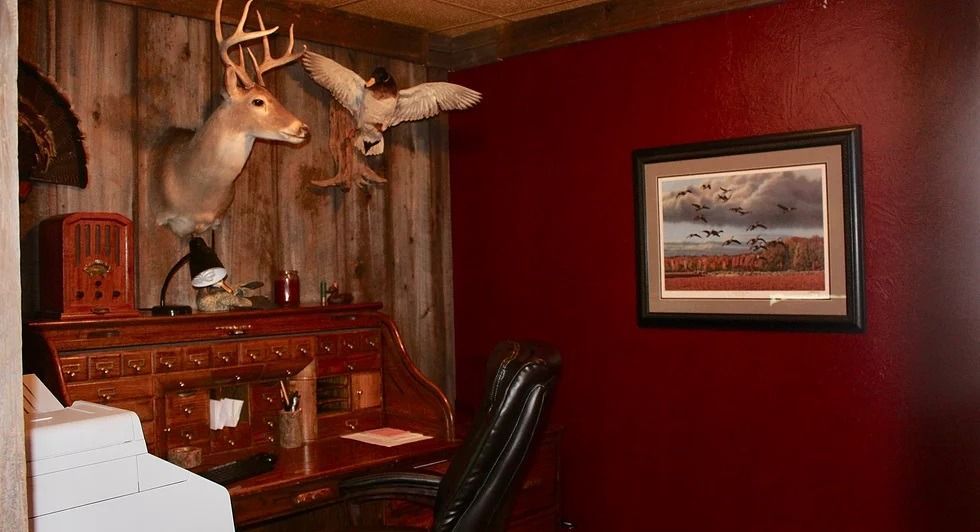 Office with wooden desk, deer and owl taxidermy, framed artwork on red wall.