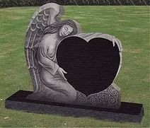Angel statue embracing heart-shaped headstone on grass. Dark stone, angel with wings.