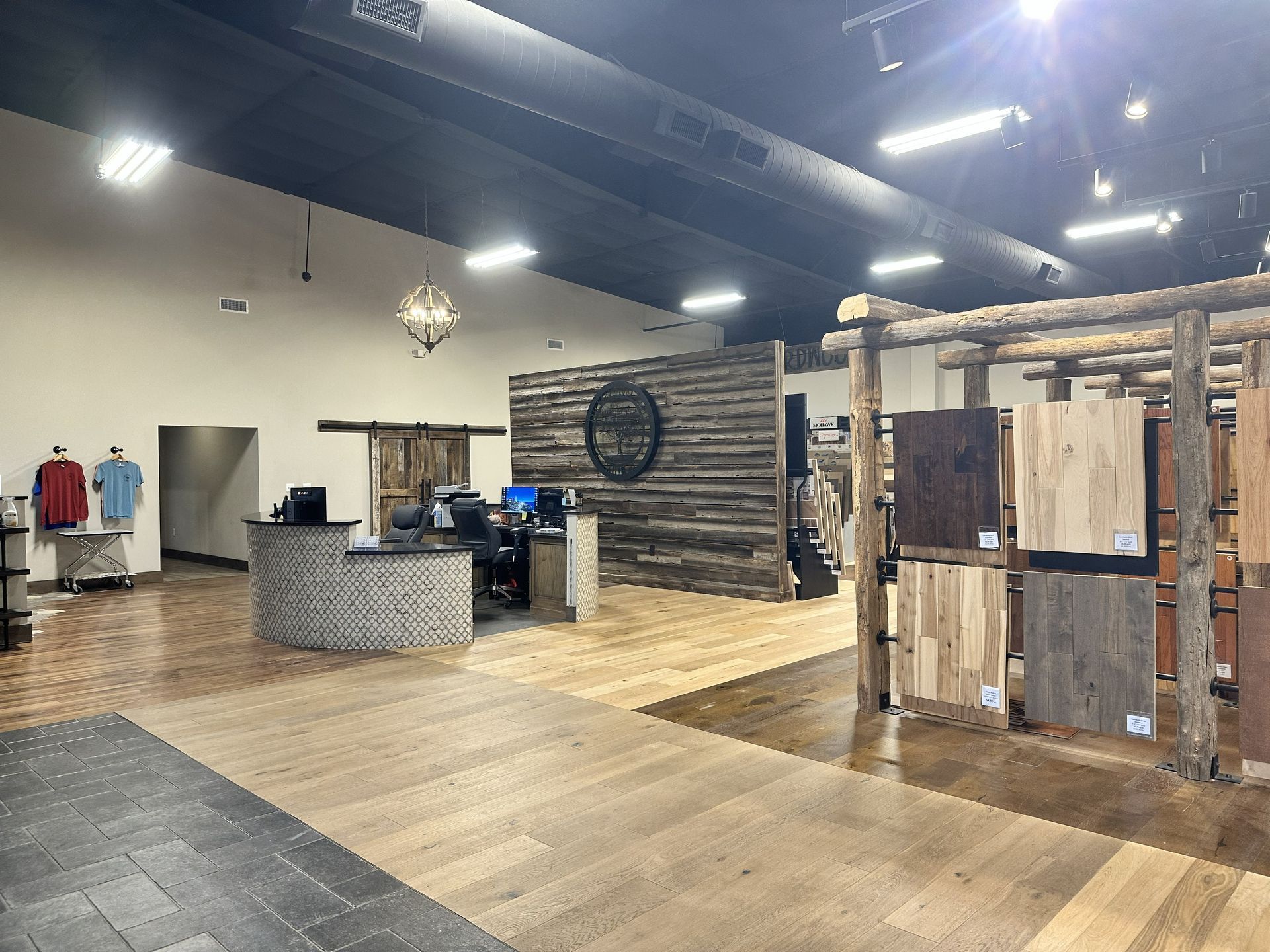 Showroom interior with a reception desk, rustic wood wall, and a wooden rack displaying various wood flooring samples.