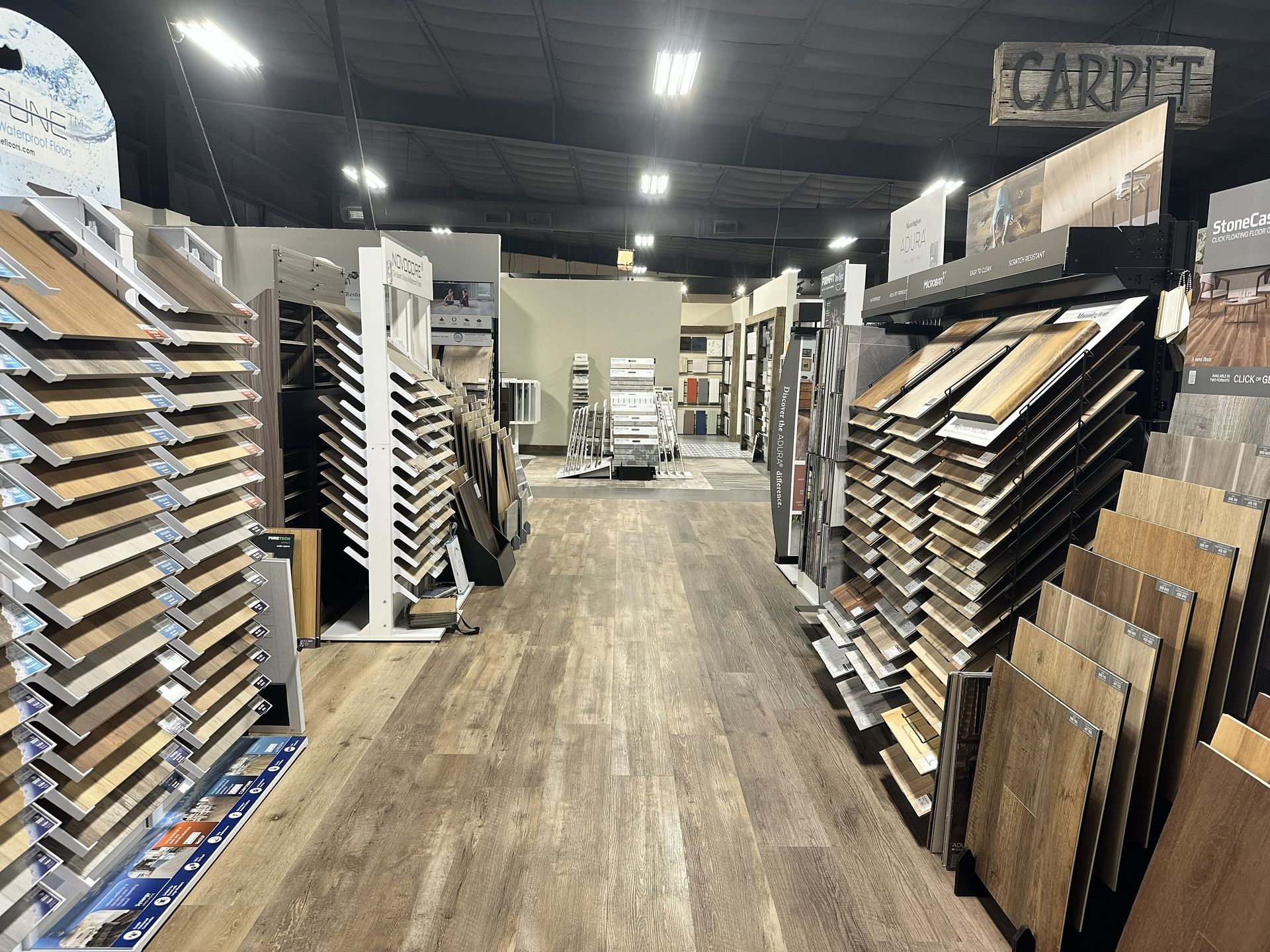 A flooring showroom aisle featuring rows of diverse wood-look and tile samples displayed on vertical racks.