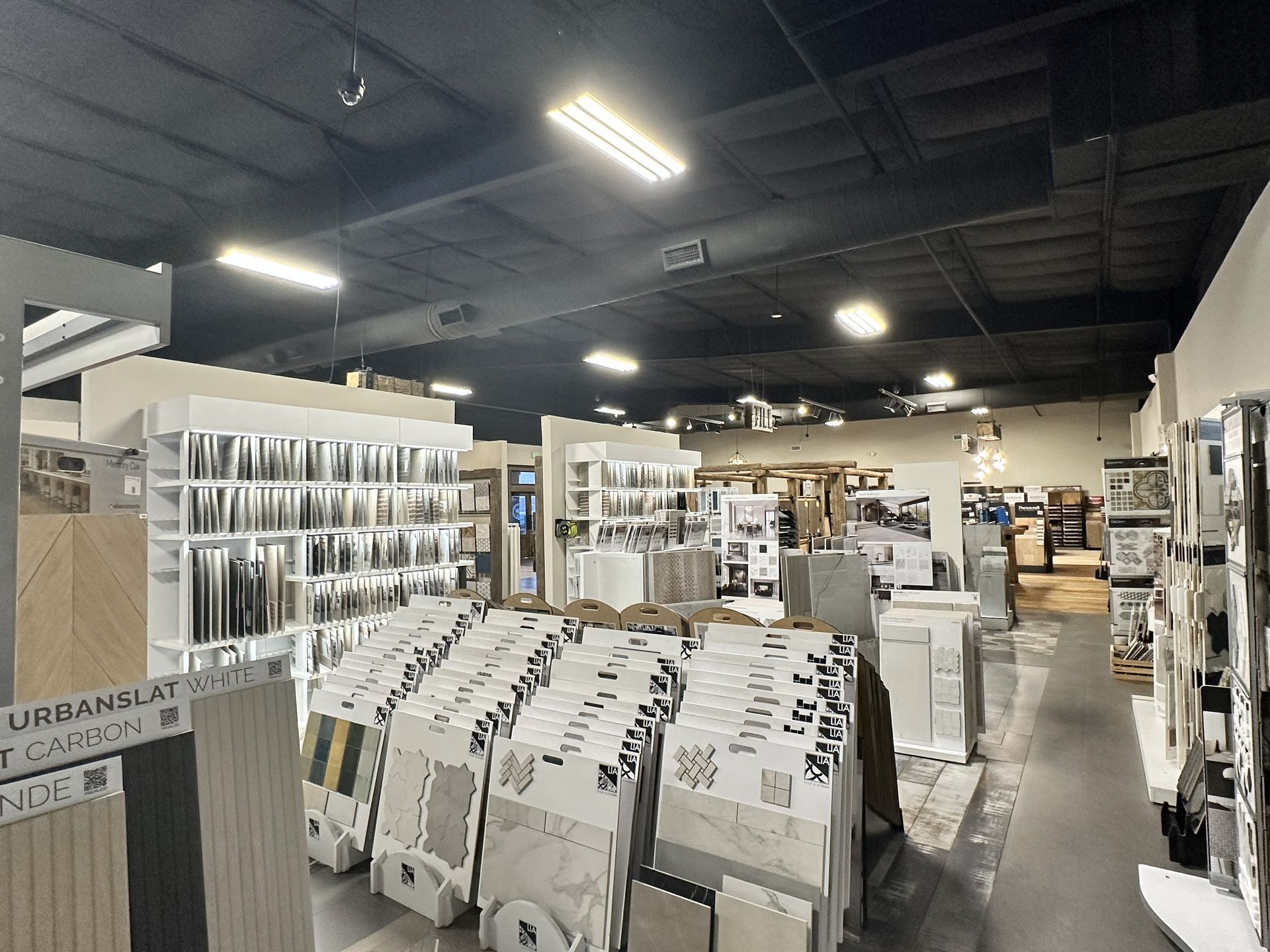 A retail showroom filled with organized tile and flooring samples displayed on racks and shelves under industrial lighting.