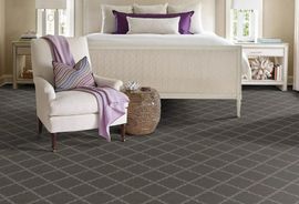 A bedroom features a white upholstered chair with a purple throw, a woven side table, and a patterned dark gray carpet.