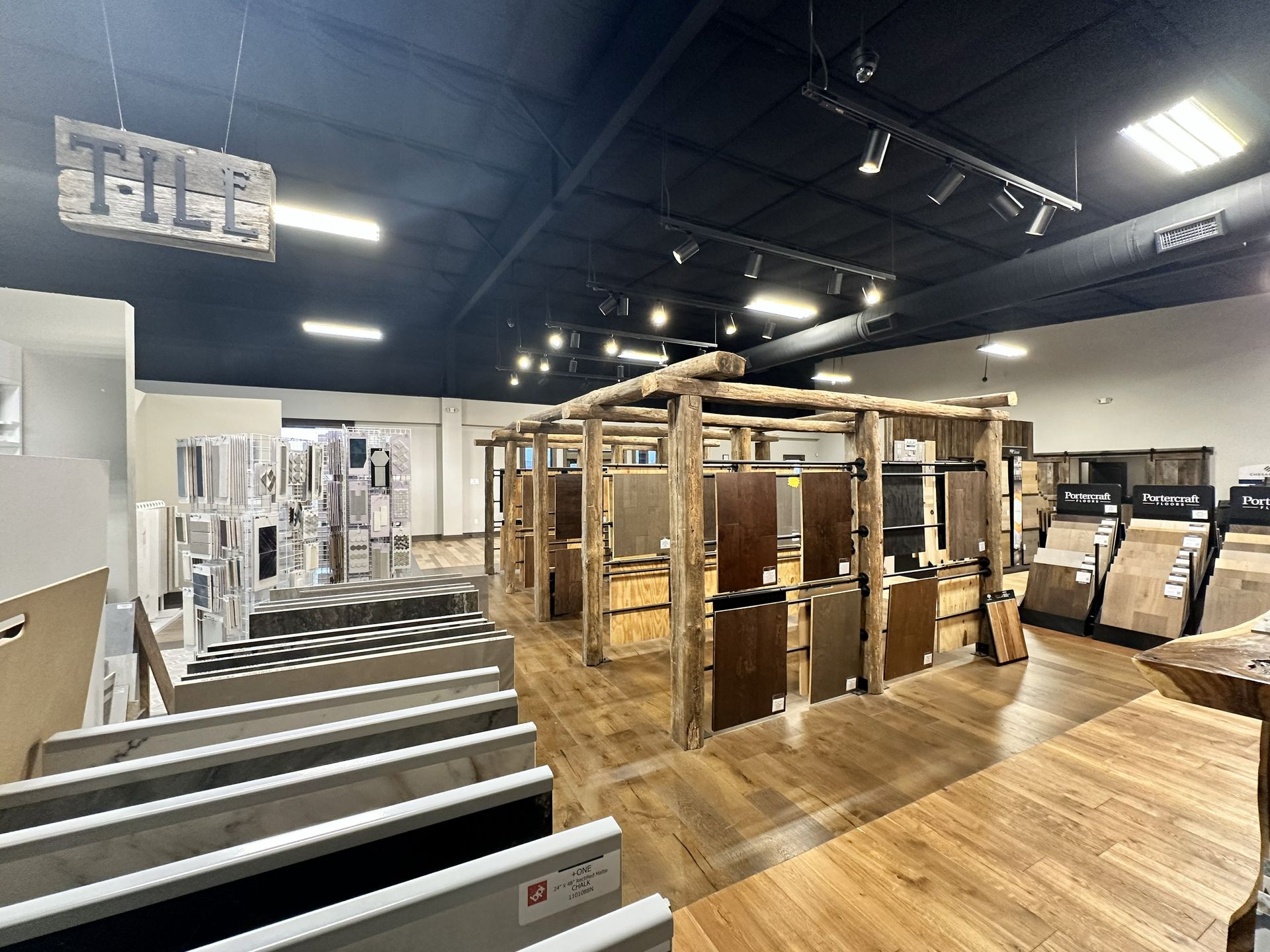 A showroom floor displays tile and flooring samples, featuring a rustic wooden structure and overhead track lighting.